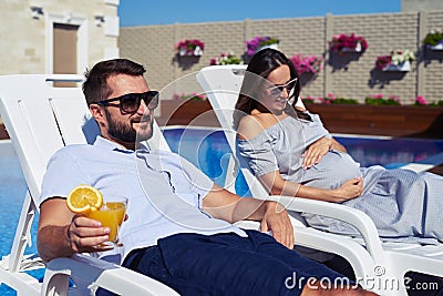 Husband and pregnant wife relaxing on lounge near pool Stock Photo