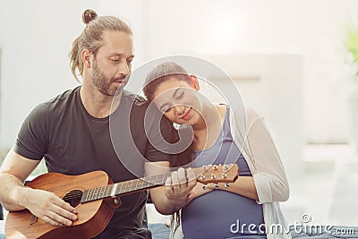 Husband with pregnant wife stay home lovely playing guitar music for baby kissing and showing love together Stock Photo