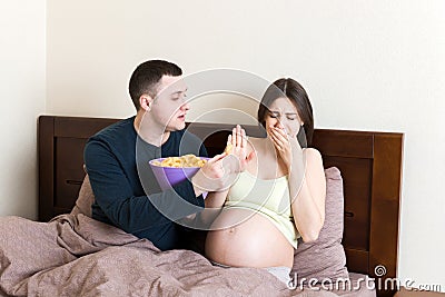 Husband tries to feed his pregnant wife with unhealthy snacks in bed but she refuses. Stop to the junk food during pregnancy Stock Photo