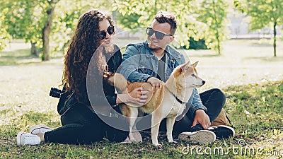 Husband and wife happy couple are fussing their pet shiba inu puppy sitting on grass in park and talking. Conversation Stock Photo