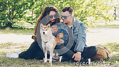 Husband and wife are using smartphone resting on lawn in park, watching screen and talking and fussing dog. Modern Stock Photo