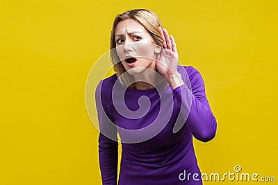 I can`t hear! Portrait of curious attentive woman keeping hand near ear. indoor studio shot isolated on yellow background Stock Photo
