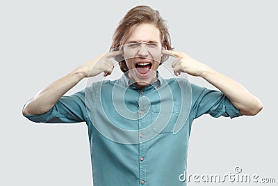 I don`t want to hear this. Portrait of angry handsome long haired blonde young man in blue shirt standing, puting fingers in ears Stock Photo