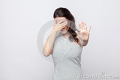 I don`t want to see this. young brunette woman with striped dress standing covering her face and showing stop gesture and try to Stock Photo