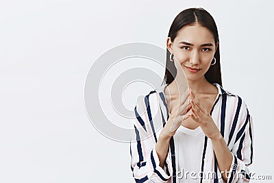 I have great evil plan. Good-looking confident and tricky woman in striped blouse, holding fingers together and smirking Stock Photo