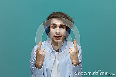 I hope. Portrait of a young beautiful man wearing white t-shirt and blue shirt in blue headphones holding his fingers crossed and Stock Photo