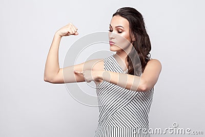 I am strong. Portrait of proud beautiful young brunette woman with makeup and striped dress standing and pointing at her biceps Stock Photo