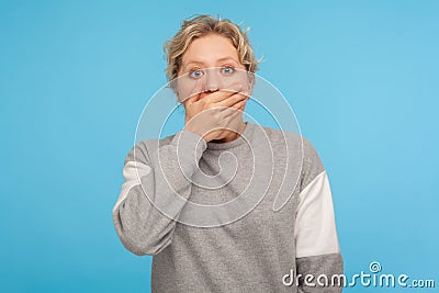 I won`t tell! Shocked intimidated woman in grey sweatshirt covering mouth with hand and looking scared at camera Stock Photo