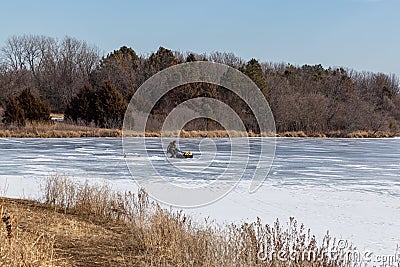 Ice fishing; frozen lake surface in winter with ice fisherman tending his stuff on the lake. Editorial Stock Photo