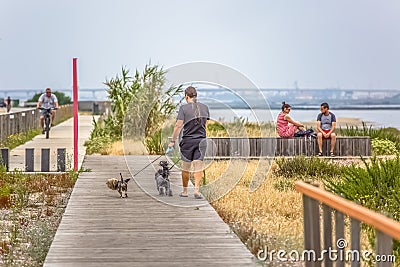 View of pedestrian path with people, man walking dogs and couple sitting on wooden bench and enjoying river, bridge view as Editorial Stock Photo
