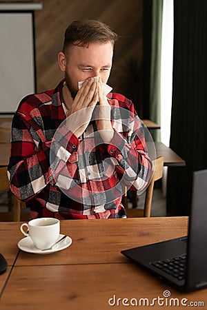 Ill caucasian man looking at laptop screen and sneezing nose in paper tissue, sitting on the office, empty space Stock Photo