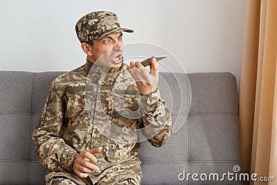 Image of aggressive military man wearing camouflage uniform sitting on sofa, soldier using voice assistant or recording voice Stock Photo