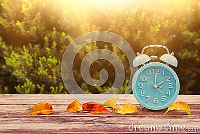 Image of autumn Time Change. Fall back concept. Dry leaves and vintage alarm Clock on wooden table outdoors at afternoon Stock Photo