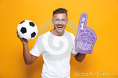 Image of beautiful man 30s in white t-shirt holding soccer ball and number one fan hand glove with finger raised while standing Stock Photo