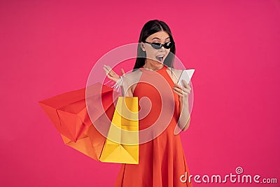 Image of a beautiful shocked young asian woman posing isolated over pink wall background holding shopping bags using mobile phone Stock Photo