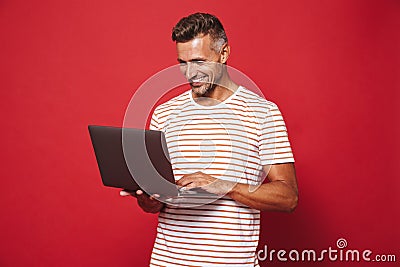 Image of brunette man in striped t-shirt smiling and holding gray laptop, isolated over red background Stock Photo