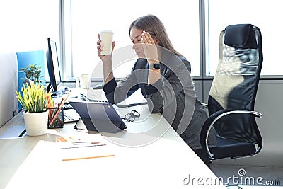 Image of business woman looking tired while sitting at her working place and holding paper cup in hand. Stock Photo