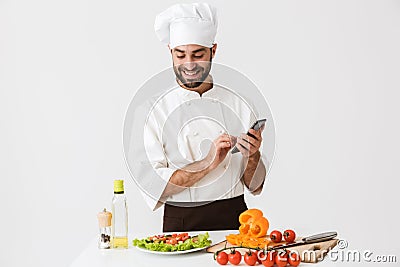 Image of caucasian chef man in uniform smiling and holding smartphone while cooking vegetable salad Stock Photo