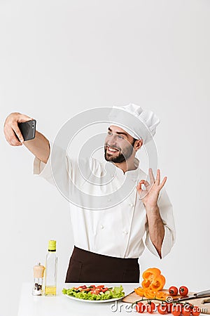 Image of caucasian cook man in uniform showing ok sign and taking selfie photo of food on smartphone at work isolated over white Stock Photo