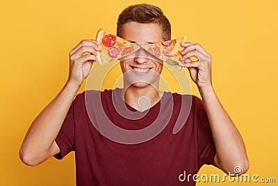 Image of cheerful handsome young boy holding two pieces of pizza in both hands, having beautiful smile, covering his eyes with Stock Photo