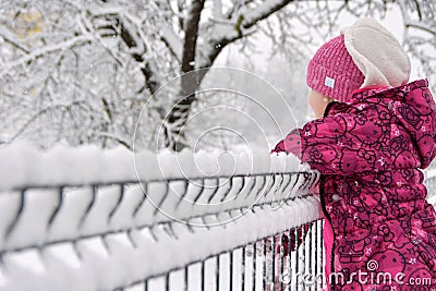 Image of child in winter hat and jacket leaning on aluminum fence with snow covered background Stock Photo