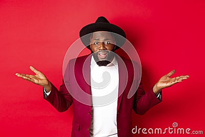 Image of confused Black man asking question, spread hands sideways and staring camera clueless, standing against red Stock Photo