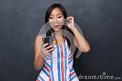 Image of delighted asian woman 30s wearing earphones using black mobile phone standing isolated Stock Photo