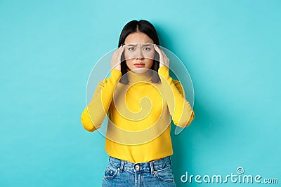 Image of distressed asian woman touching head and frowning, feeling headache, standing troubled against blue background Stock Photo