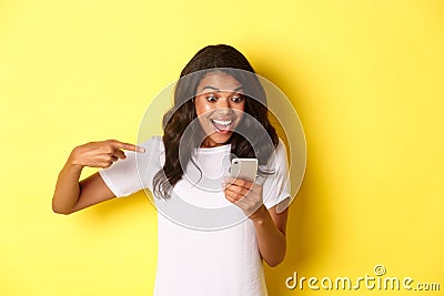 Image of excited african-american girl, looking and pointing finger at mobile phone, seeing something amazing online Stock Photo