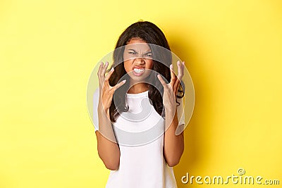 Image of frustrated and angry african-american girl, grimacing and shaking hands mad, standing over yellow background Stock Photo