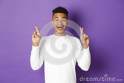 Image of handsome african-american young guy in white sweatshirt, looking hopeful and smiling with crossed fingers Stock Photo