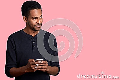 Image of handsome guy with dark skin, in black shirt, stands and holds phone in hand, looks aside with uncomprehending, hears Stock Photo