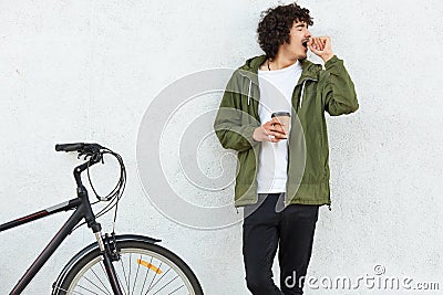 Image of handsome male yawns as wants to sleep, keeps hands near mouth, drinks strong hot coffee to feel refreshed, being tired af Stock Photo