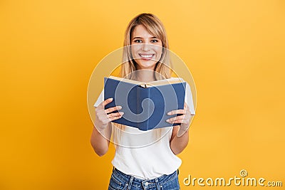 Cheerful emotional young blonde woman posing isolated over yellow wall background dressed in white casual t-shirt holding book Stock Photo