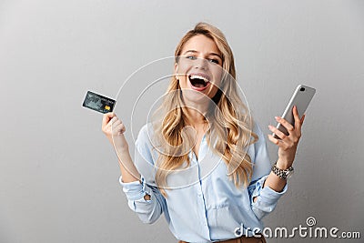 Happy young pretty blonde business woman posing isolated grey wall background holding credit card using mobile phone Stock Photo