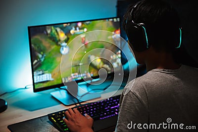 Image of immersed teenage gamer boy playing video games on computer in dark room, wearing headphones and using backlit colorful k Stock Photo