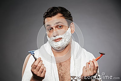 Image of indecisive man with shaving foam on his face holding two razors, isolated over gray background Stock Photo