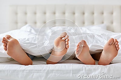 Image of man and woman bare feet under blanket in bedroom. Unrecognizable husband and wife spend free time in comfortable bed, enj Stock Photo