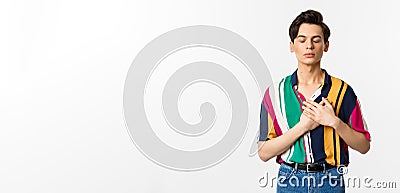 Image of nostalgic young gay man standing with closed eyes and hands on heart, remember something, standing over white Stock Photo