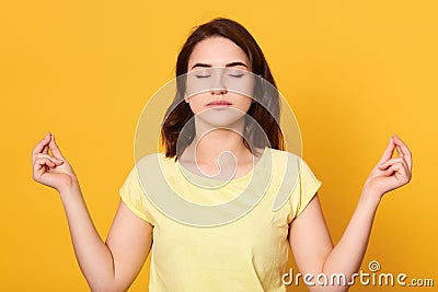 Image of peaceful good looking brunette wearing yellow t shirt, raising hands, having meditation, closing eyes, being alone, Stock Photo