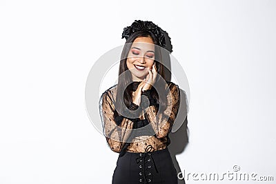 Image of sensual, beautiful asian woman in gothic lace dress and black wreath smiling coquettish, touching face and Stock Photo