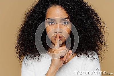 Image of serious confident dark skinned woman makes shush gesture, keeps fore finger over lips, tells secret information, asks not Stock Photo