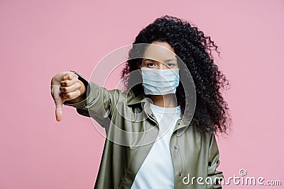 Image of serious curly haired young woman wears medical mask, keeps thumb down, shows dislike gesture, avoids spreading of corona Stock Photo