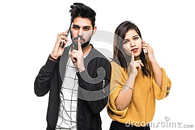 Image of serious young loving indian couple talking by mobile phones make silence gesture over white wall background Stock Photo