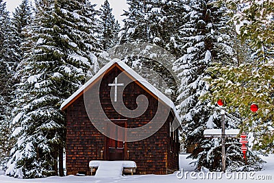 Image of a small rural church sitting amongst snow covered spruce trees, one which has a few red Christmas ornaments on it. Stock Photo