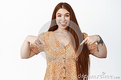 Image of smiling redhead woman pointing fingers down, showing event announcement, advertising store, standing over white Stock Photo