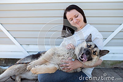 Image of smiling woman in jeans and white jacket sitting next to happy shepherd puppy against white wooden building Stock Photo