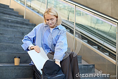 Image of smiling young woman, student packing her laptop in backpack, sitting on public stairs with cup of coffee, hurry Stock Photo