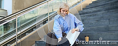 Image of smiling young woman, student packing her laptop in backpack, sitting on public stairs with cup of coffee, hurry Stock Photo