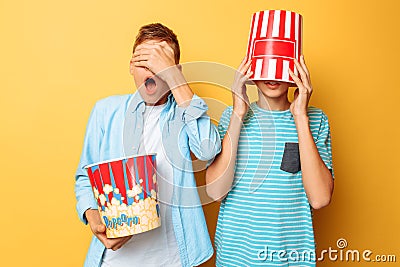 Image of two frightened teenagers, guys watching a horror movie and hiding behind a bucket of popcorn on a yellow background Stock Photo